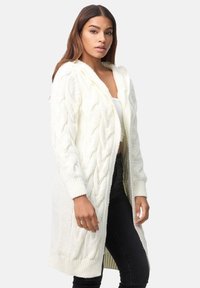 Cardigan long en tricot torsadé de couleur crème avec une capuche, doté de poignets et d'un ourlet côtelés. Porté par-dessus un haut ajusté et un jean noir.