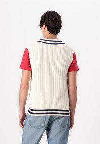 Kremfarget ribbestrikket vest med marineblå stripet krage og bunnband, båret over en rød T-skjorte, kombinert med lyseblå jeans.