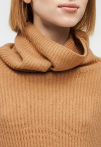 Πουλόβερ με λαιμόκοψη τύπου καψούλι σε ζεστό, απαλό καφέ χρώμα με ραβδωτή υφή. Η κοντινή λήψη αναδεικνύει το μαλακό υλικό και τη χαλαρή εφαρμογή γύρω από το λαιμό.