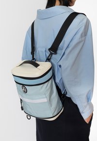 Ljusblå ryggsäck med ett handtag på toppen, justerbara svarta remmar och ett vitt dragkedjestopp. Har en frampocket med ett vitt rand.