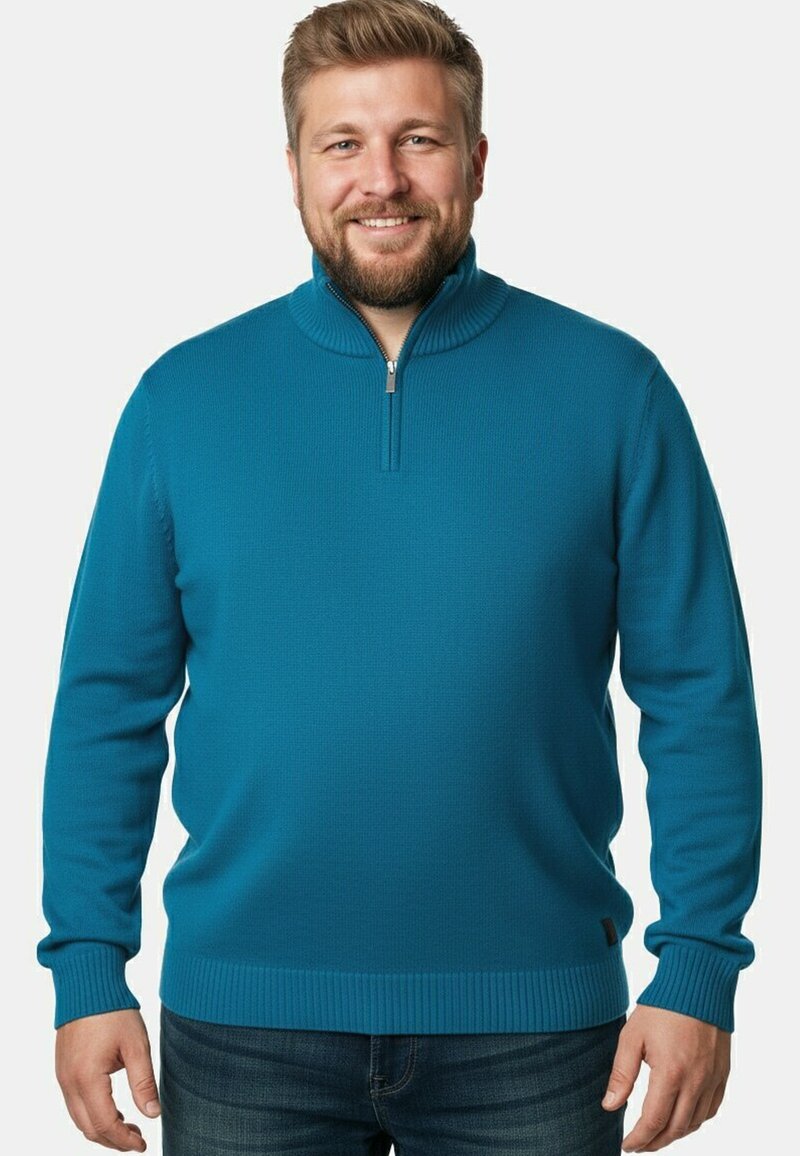 Pull à col zippé bleu sarcelle avec une texture côtelée, manches longues et ourlet ajusté. Porté avec un jean bleu, vu de face.