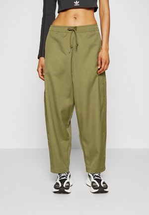 Femme portant un pantalon large kaki avec des boutons sur le côté, un crop top noir avec le logo Adidas, et des baskets noir et blanc, debout devant un fond blanc.