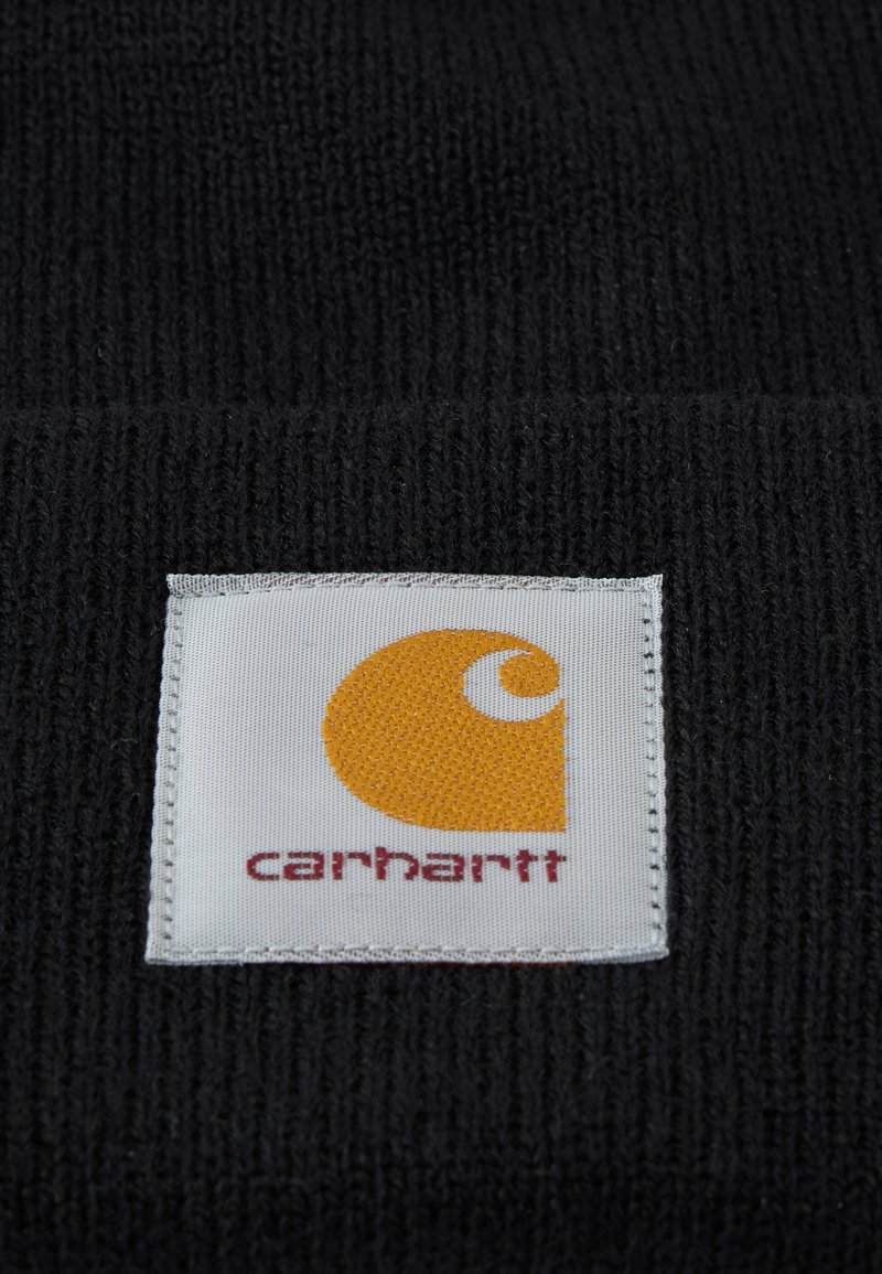 Etiqueta cuadrada de tela blanca con un logo curvado naranja y texto rojo "carhartt" cosido sobre tela negra de punto.