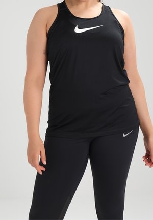 Osoba ubrana w czarny bezrękawnik Nike i czarne legginsy Nike, stojąca na jednolitym jasnoszarym tle.