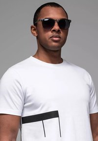 Hvid t-shirt med et sort vandret stribemønster på den nederste front. Modellen bærer mørke solbriller.