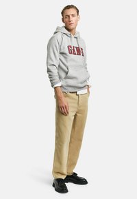 Felpa grigia in cotone con logo "GANT", pantaloni beige e scarpe nere. Il modello si trova in una posa rilassata su sfondo bianco.
