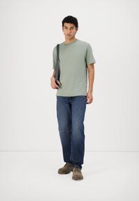 Light green short-sleeve t-shirt, relaxed fit, paired with blue denim jeans. The model wears brown footwear and carries a black strap bag.