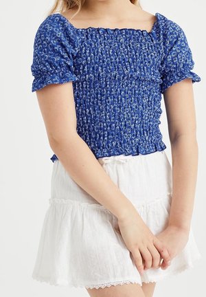 Haut bleu smocké avec manches bouffantes arborant un motif floral, associé à une jupe blanche à volants avec bordure en dentelle et tissu texturé.