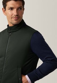 Giacca senza maniche verde scuro con colletto alto, zip frontale, tasche laterali e maniche di maglione blu navy. Tessuto dalla consistenza liscia.