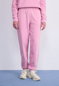 Pantalons de survêtement rose clair avec une taille élastique et des poignets, comportant des poches latérales et un détail de logo subtil. Portés avec des chaussures de sport beiges.