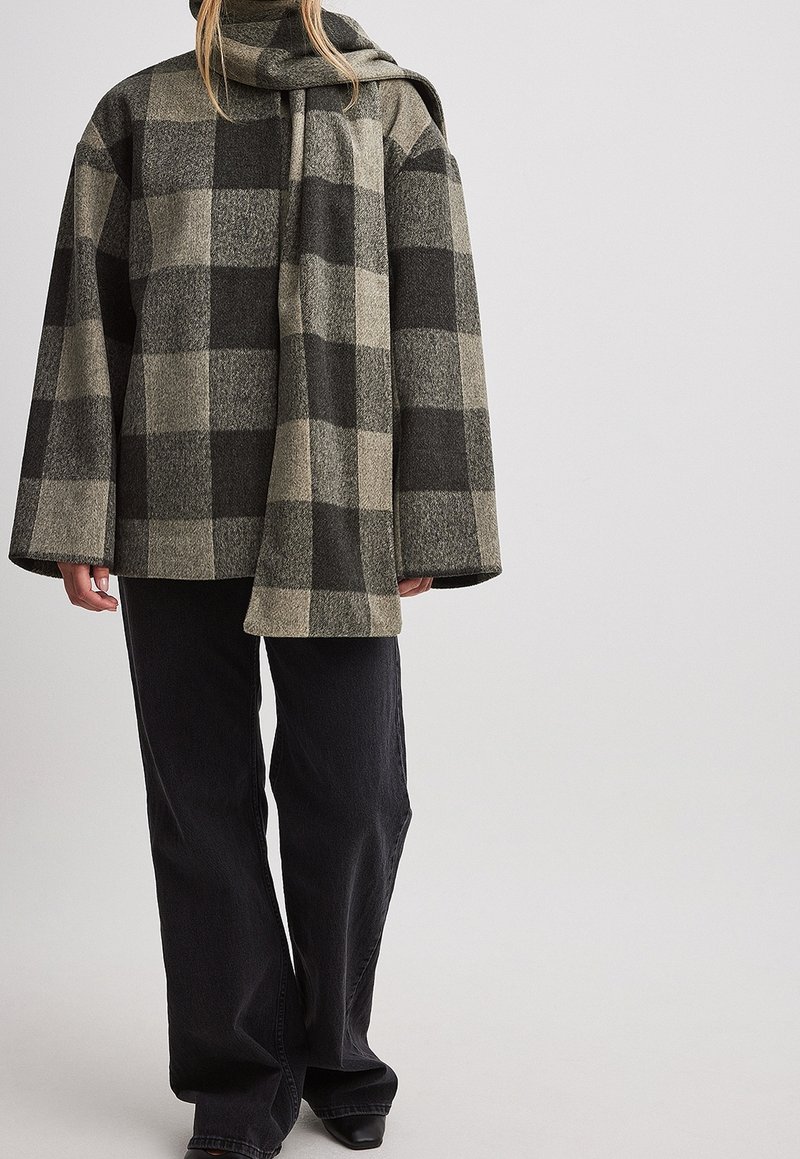 Modellen bär en lös, oversized rutig yttre jacka i jordnära färger, tillsammans med svarta vida byxor och eleganta svarta skor.