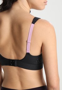 Svart sports-bh med rosa stropper, en jevn tekstur og krok-og-øyne lukking. Har subtile mønstrede detaljer på baksiden.