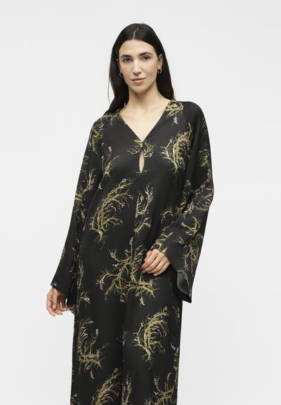 Γυναίκα με μακριά σκούρα μαλλιά που φοράει ένα άνετο μαύρο φόρεμα με πράσινα και ροζ φυτικά μοτίβα και φαρδιά μανίκια σε λευκό φόντο.