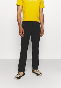 Zwarte outdoorbroek met een slim fit, voorzien van ritszakken en versterkte stiksels, gecombineerd met een gele t-shirt en gestreepte schoenen.