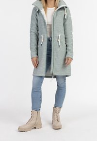 Ljusgrön pufferjacka med hög krage, dragkedjeförslutning, sidfickor och snoddar. Bärs med ljusblå jeans och beiga kängor.