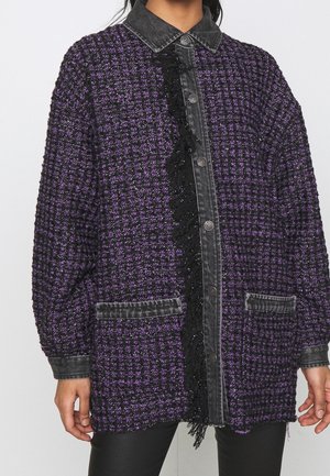 Femme portant une veste texturée violette et noire avec fermeture à boutons sur le devant, col et poignets noirs, et deux poches avant.
