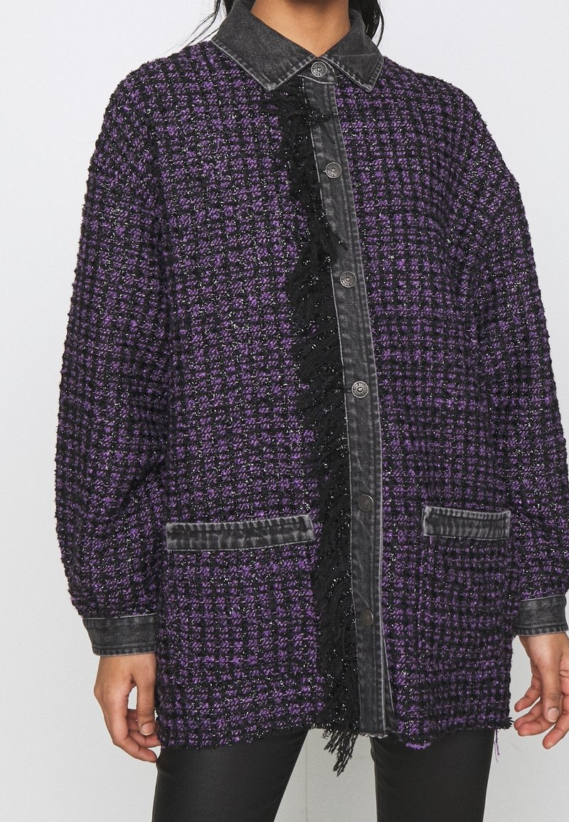 Femme portant une veste texturée violette et noire avec fermeture à boutons sur le devant, col et poignets noirs, et deux poches avant.