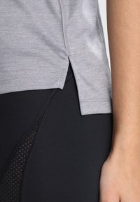 Camisa de manga corta gris claro con aberturas laterales, combinada con leggings negros que presentan un panel de malla texturizada en la parte inferior de la pierna.