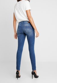 Jeans ajustados en denim azul oscuro con una textura suave, diseño de cinco bolsillos y detalles de costura sutiles. Combinados con tacones altos negros.