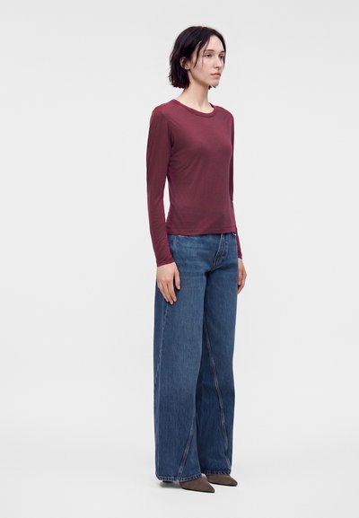 Femme debout, face à droite, portant une chemise bordeaux à manches longues, un jean large bleu et des chaussures marron pointues sur un fond clair uni.