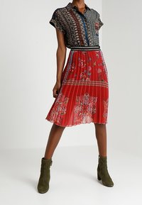 Chemise à manches courtes à motifs dans des tons foncés, associée à une jupe plissée rouge ornée d'imprimés floraux. Des bottines vert olive complètent la tenue.