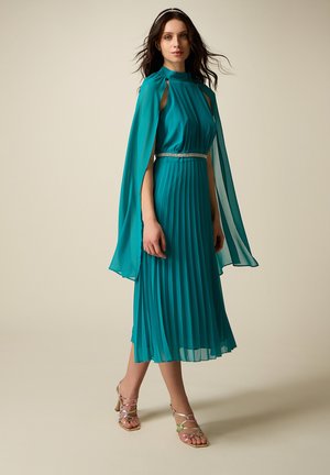 Donna che indossa un abito midi plissettato color teal con maniche a mantella, una cintura gioiello, tacchi metallici con cinturini e una fascia per capelli, in piedi su uno sfondo beige.