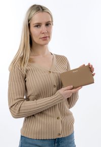 Pitkät vaaleat hiukset omaava nainen beige ribbineuleinen neuletakki yllä pitää käsissään hailakanruskeaa vetoketjullista lompakkoa, jossa on logo valkoista taustaa vasten.