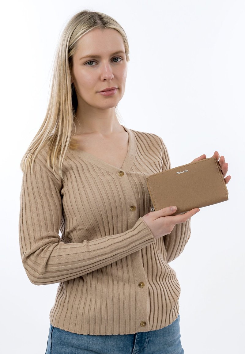 Pitkät vaaleat hiukset omaava nainen beige ribbineuleinen neuletakki yllä pitää käsissään hailakanruskeaa vetoketjullista lompakkoa, jossa on logo valkoista taustaa vasten.