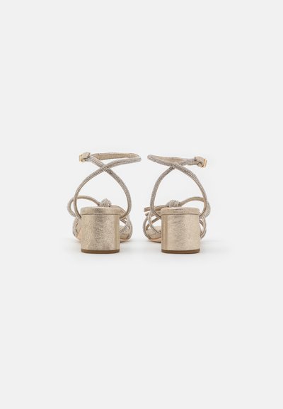 Sandales à talons blocs métalliques dorés avec des brides entrecroisées, ornées de petites perles, dotées d'une finition lisse et texturée.