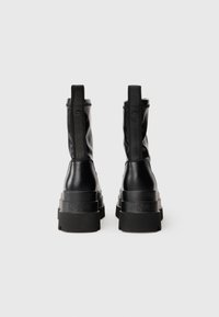 Botas negras de tobillo con un acabado de cuero suave, puntera redonda, suela de plataforma gruesa y una marca prominente en el talón trasero y en la correa.