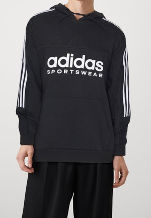 Person trägt schwarzen Adidas-Sportpullover mit weißem Logo und drei Streifen an den Ärmeln, kombiniert mit lockeren schwarzen Hosen.
