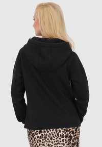 Felpa nera con cappuccio, dal texture morbido, caratterizzata da un fit rilassato e cuciture visibili. Il modello la indossa sopra un pantalone con stampa leopardata.
