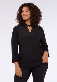 Blusa nera con scollatura a chiave e colletto decorativo con perline. Maniche lunghe con leggero svaso e tessuto strutturato.
