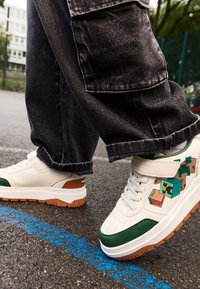 Baskets blanches avec des accents verts et une semelle marron, ornées d'un motif graphique de blocs colorés. Portées avec un jean foncé et ample.