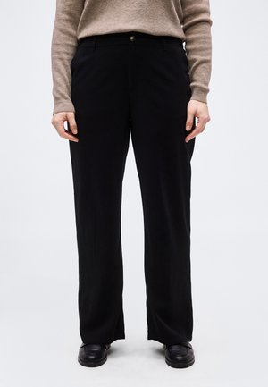 Fekete bő szárú nadrágot, fekete cipőt és bézs hosszú ujjú pulóvert viselő személy áll sima világos háttér előtt.