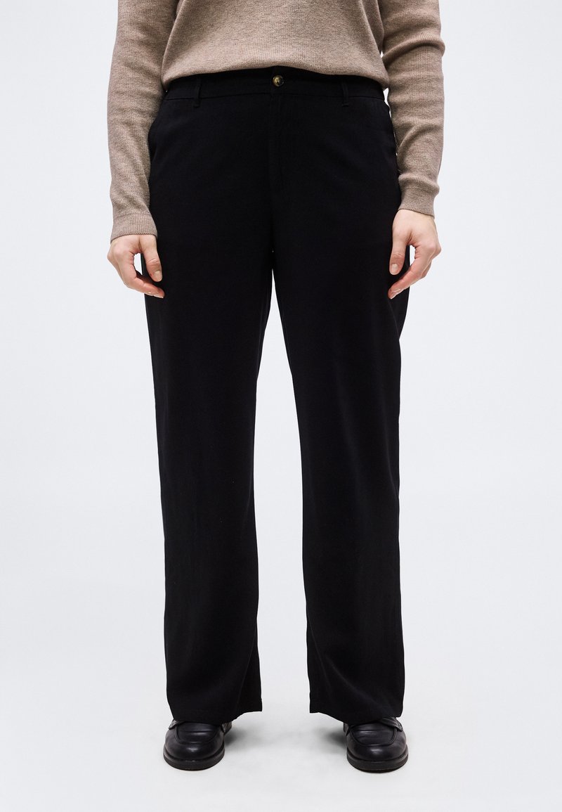 Άτομο που φοράει μαύρο παντελόνι με φαρδιά μπατζάκια, μαύρα παπούτσια και μπεζ πουλόβερ με μακριά μανίκια, στέκεται μπροστά σε ένα απλό ανοιχτό φόντο.