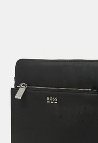 Pochette nera in materiale sintetico con superficie testurizzata, tasca frontale con zip e hardware in argento. Logo del marchio "BOSS" in evidenza.