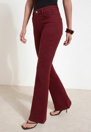 Woman wearing burgundy high-waist flare jeans, black tank top, brown bangle bracelets, and black open-toe heeled sandals, standing on beige floor.