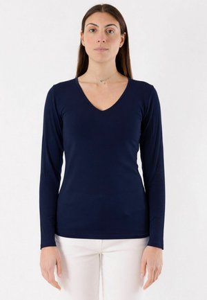 Giovane donna in piedi, rivolta verso l'obiettivo, che indossa una maglietta a maniche lunghe con scollo a V blu navy e pantaloni beige chiaro su sfondo bianco.