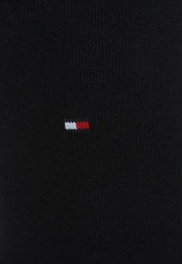 Svarta räfflade strumpor med små, broderade horisontella ränder i rött, vitt och blått nära toppen. Mjuk textur och formar sig efter foten.