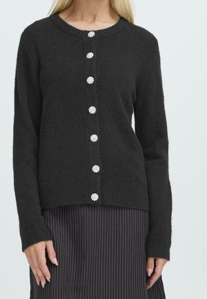 Femme portant un cardigan noir en tissu duveteux avec six boutons décoratifs en forme de fleurs argentées et une jupe noire à fines rayures.