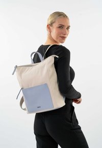 Mochila de tela beige con un bolsillo frontal azul claro. Cuenta con doble tira y un cierre con cremallera. Diseño simple y funcional.