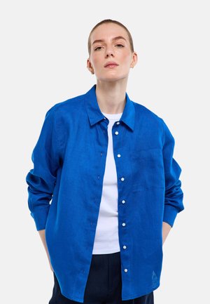 Femme à la peau claire et aux cheveux tirés en arrière, portant une chemise boutonnée bleu vif par-dessus un haut blanc, debout les mains dans les poches, fond neutre.