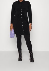 Una persona indossa un vestito nero con bottoni e arricciature, leggings neri, stivaletti neri chunky con lacci e tiene in mano una piccola borsa a tracolla viola con texture.