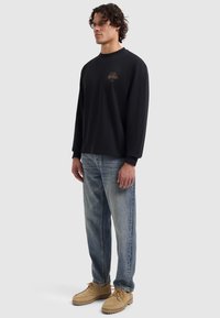 Schwarzes Langarmshirt mit einem kleinen, goldenen Logo auf der Brust, kombiniert mit ausgewaschenen blauen Jeans und hellbeigen Schuhen. Lässiger Schnitt und Design.