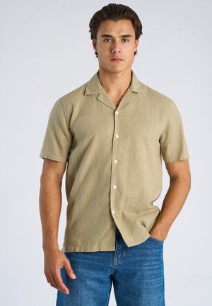 Mann mit mittlerem Hautton, der ein beigefarbenes, kurzärmeliges Hemd mit Knöpfen und blaue Jeans trägt, steht mit einer Hand in der Hosentasche.