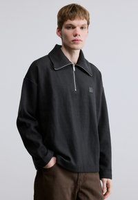 Pull noir à manches longues avec col montant et fermeture à demi-fermeture éclair, doté d'un petit patch logo et d'une coupe décontractée. Le tissu semble doux et texturé.