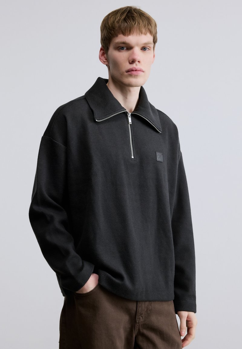 Pull noir à manches longues avec col montant et fermeture à demi-fermeture éclair, doté d'un petit patch logo et d'une coupe décontractée. Le tissu semble doux et texturé.