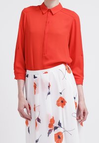 Rote Bluse mit Kragen und langen Ärmeln, mit einer Knopfleiste versehen, kombiniert mit einem weißen Rock im Blumenmuster, der orangefarbene und marineblaue Akzente aufweist.