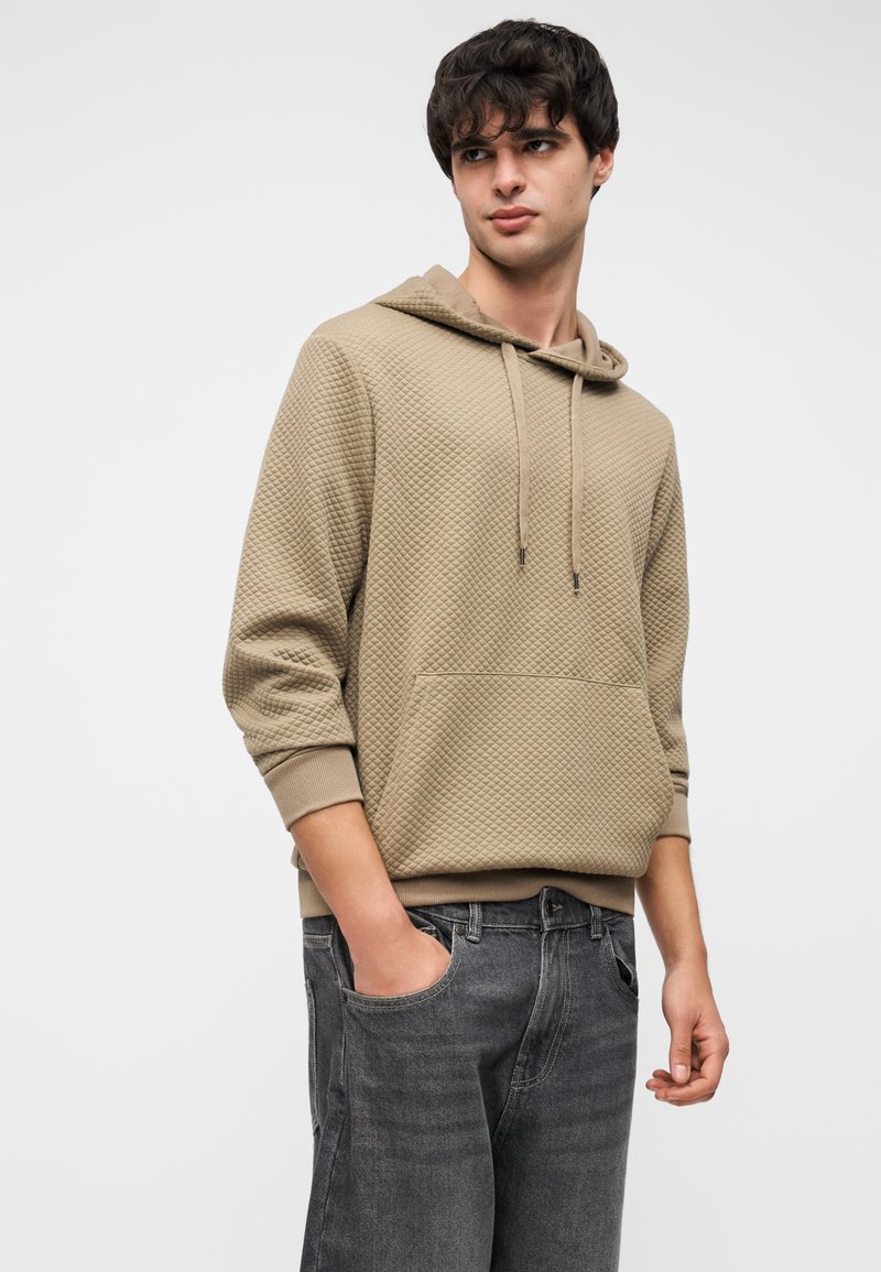Sweat à capuche beige avec un motif texturé, poche avant et cordons de serrage, associé à un jean gris foncé. Palette de couleurs neutres.